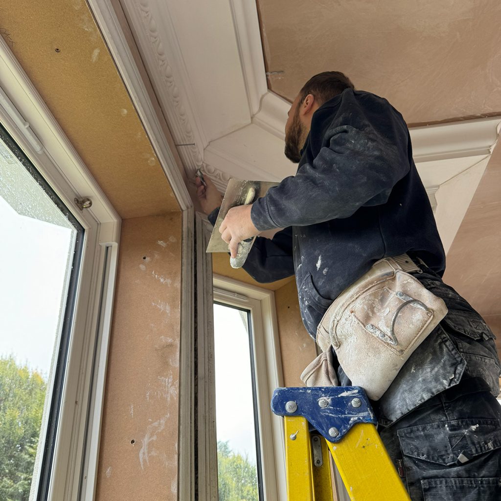 Plaster cornice installation in progress with tradesman fitting moulding at ceiling level on ladder